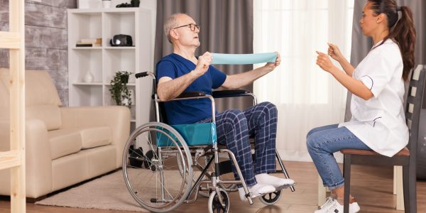 Senior man with disability in wheelchair doing recovery exercise with resistance band. Disabled handicapped old person with social worker in recovery support therapy physiotherapy healthcare system nursing retirement home